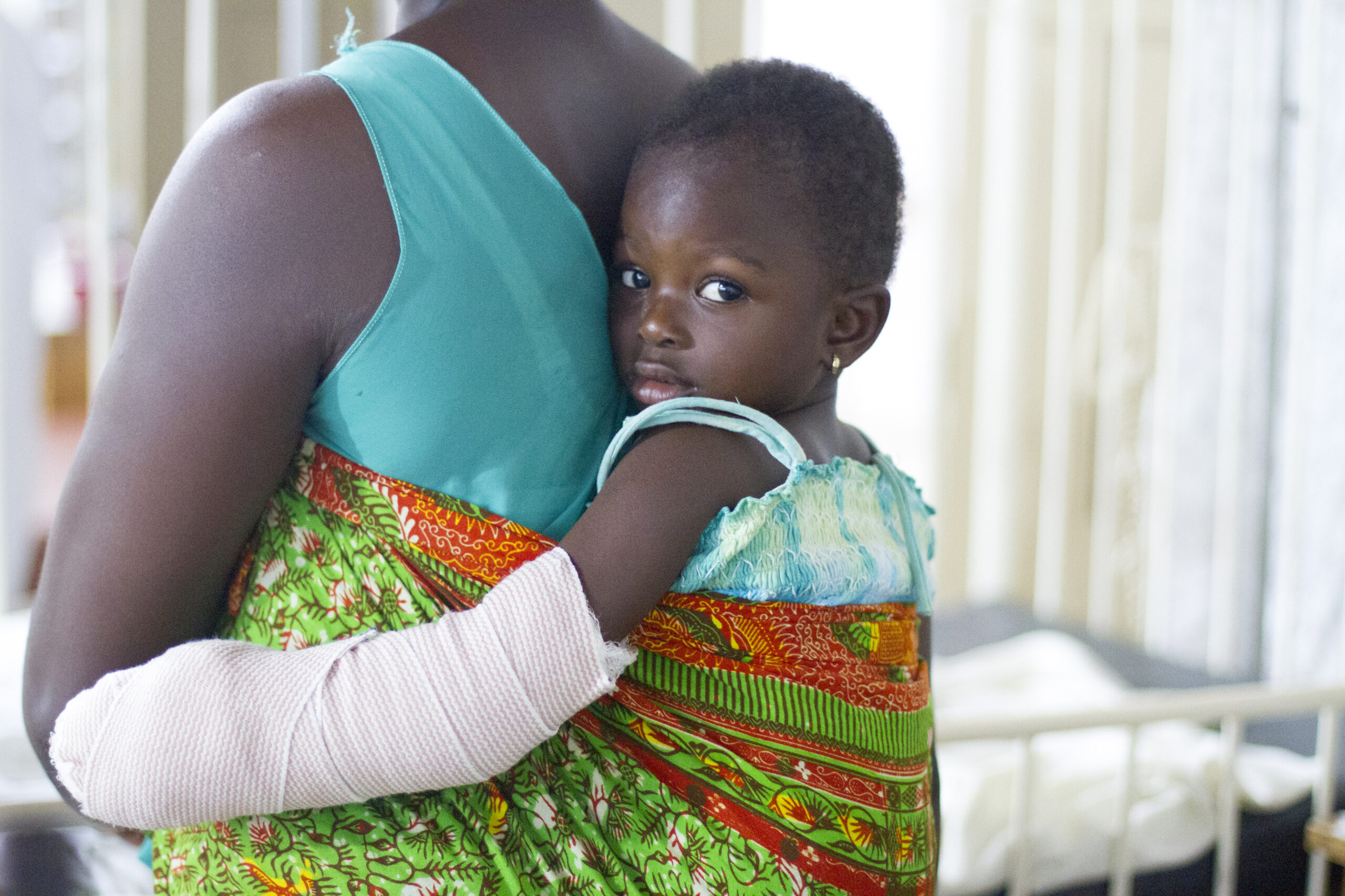 A young patient looks back over her mother's shoulder, arm bandaged after care